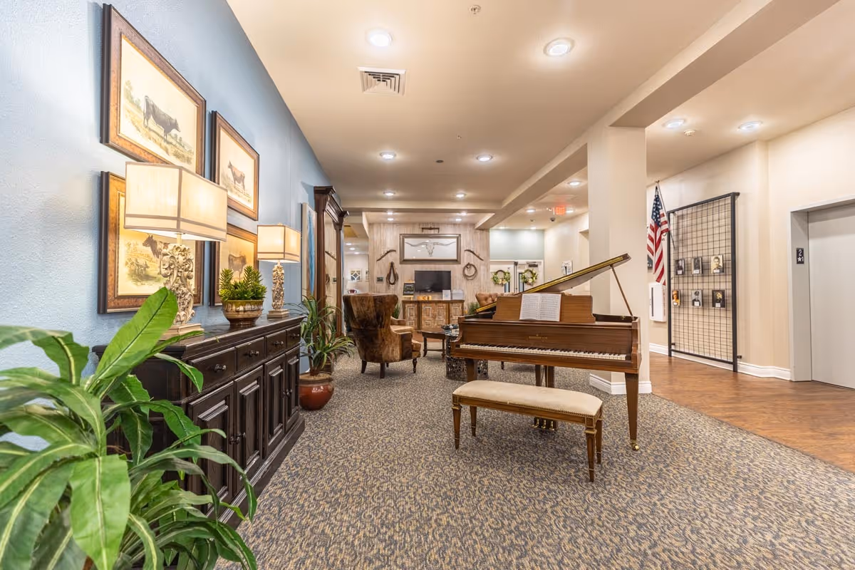Interior view of a senior living facility common area featuring a grand piano with a bench, a brown upholstered armchair, a dark wooden sideboard with lamps and framed pictures of cows on the wall, potted plants, and a television mounted on a wooden cabinet. The area has carpeted flooring, recessed ceiling lights, and an American flag near an elevator.