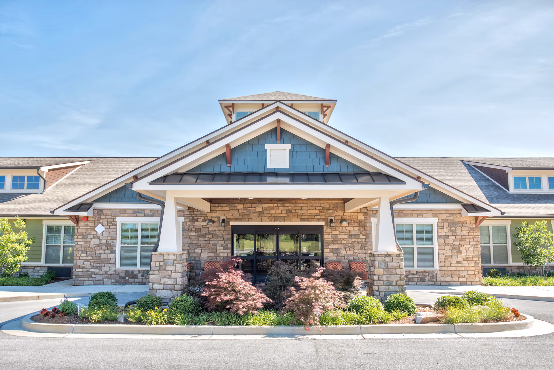 Front entrance of a senior living building with a covered porte-cochère, stone facade, and landscaped plants.