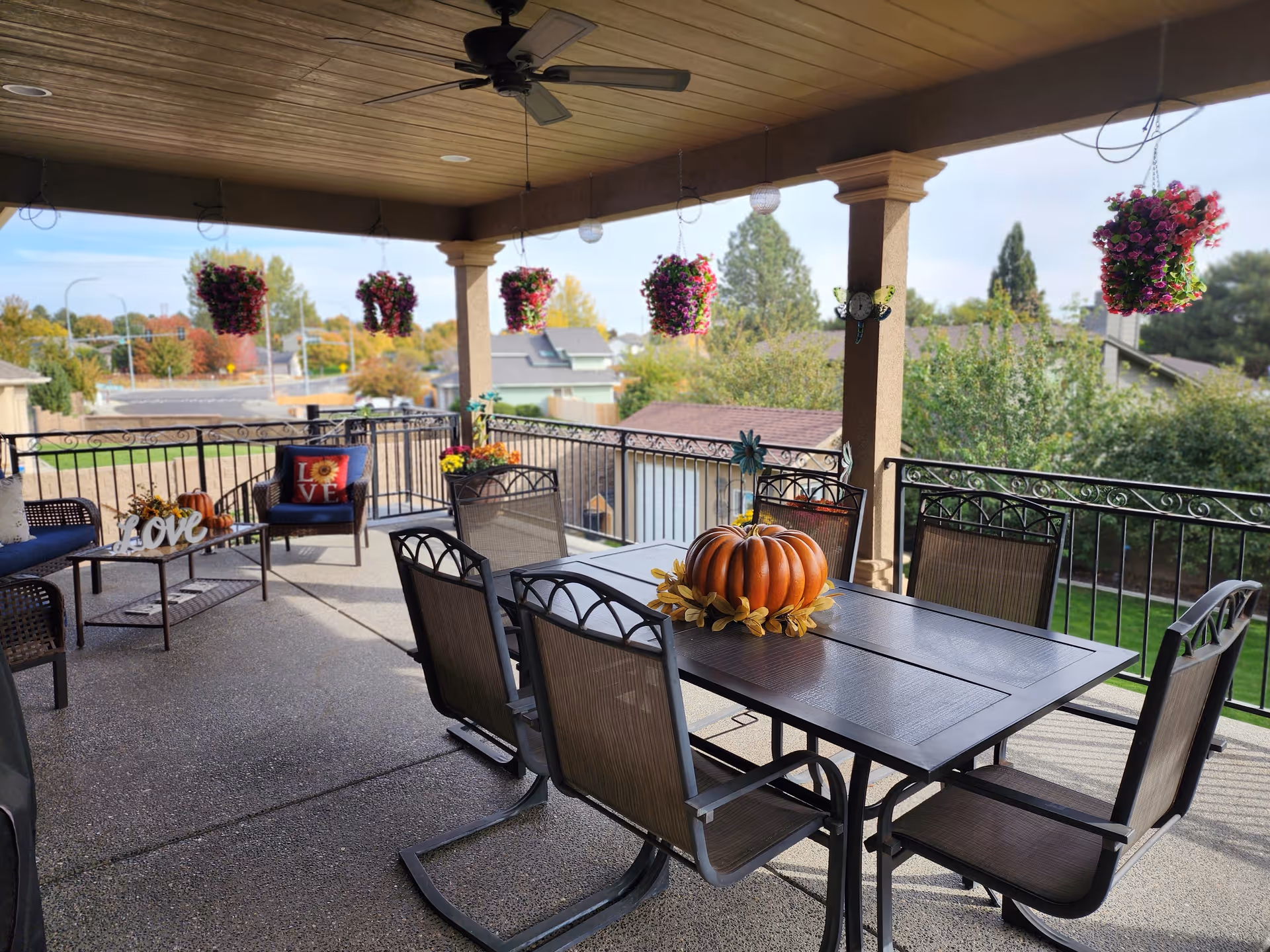 Covered outdoor patio area with a dining table and six chairs. The table has a decorative pumpkin centerpiece. Hanging flower baskets are suspended from the ceiling. There is additional seating with a small table and chairs, decorated with pillows and a 'love' sign. The patio overlooks a neighborhood with trees and houses.