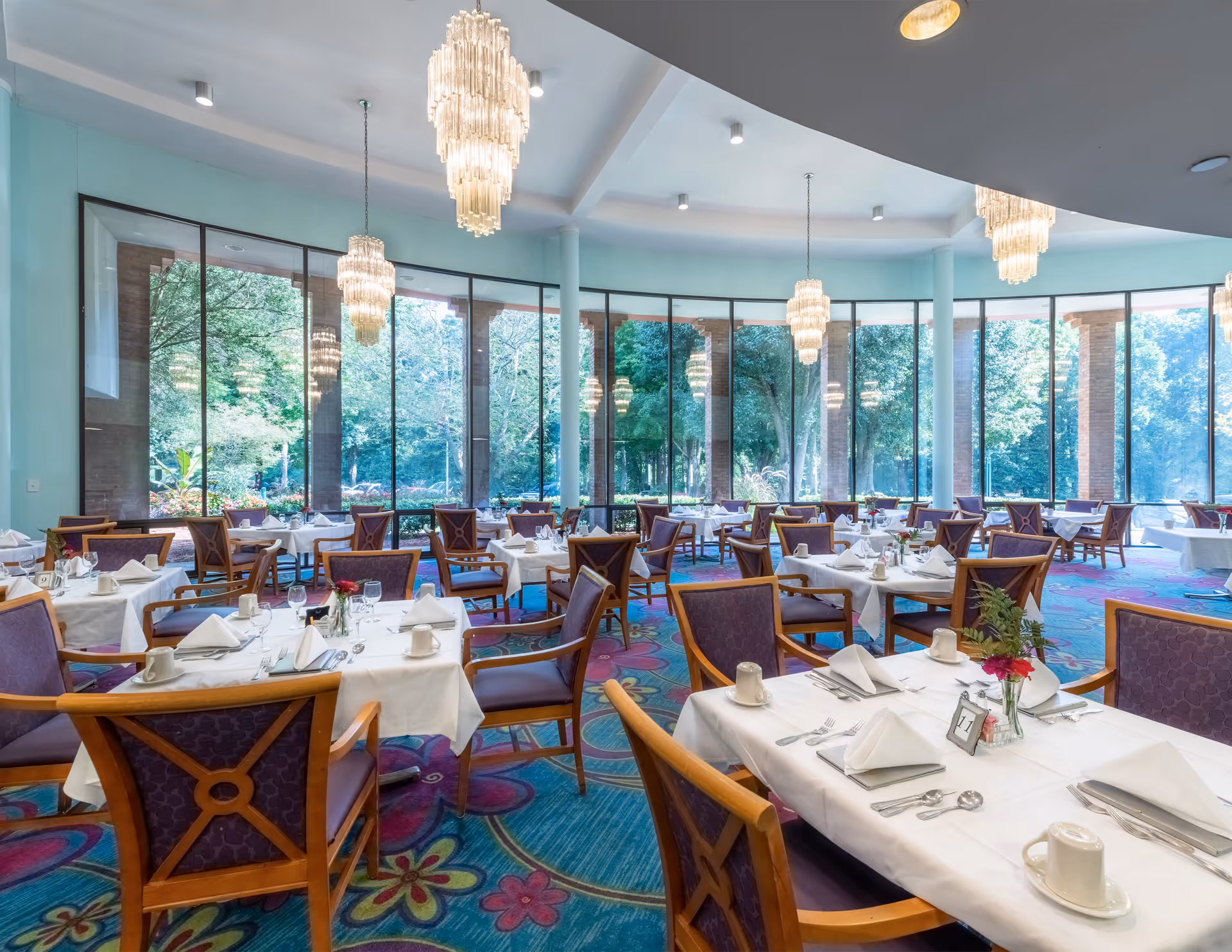 A bright dining room with large floor-to-ceiling windows offering a view of green trees outside. The room has multiple tables covered with white tablecloths, each set with white napkins, cups, glasses, and silverware. Wooden chairs with purple cushions surround the tables. Elegant chandeliers hang from the ceiling, and the carpet features a colorful floral pattern.