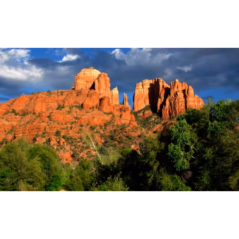 Red sandstone rock formations rising above green trees under a dramatic cloudy blue sky.