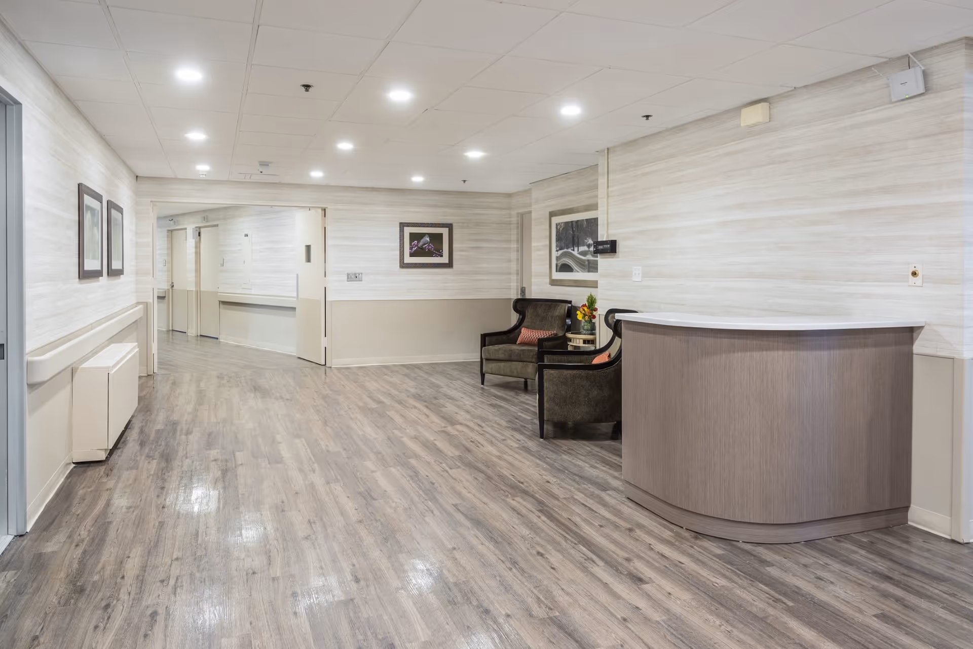 Spacious, well-lit hallway lobby with a curved reception desk, two armchairs and side table, framed art, and wood-look flooring.