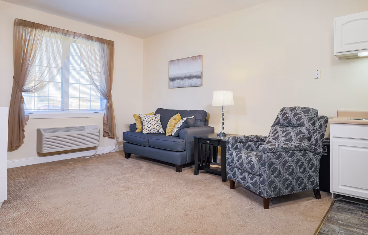 Small furnished living area with a sofa, patterned armchair, side table with lamp, and a window with curtains.