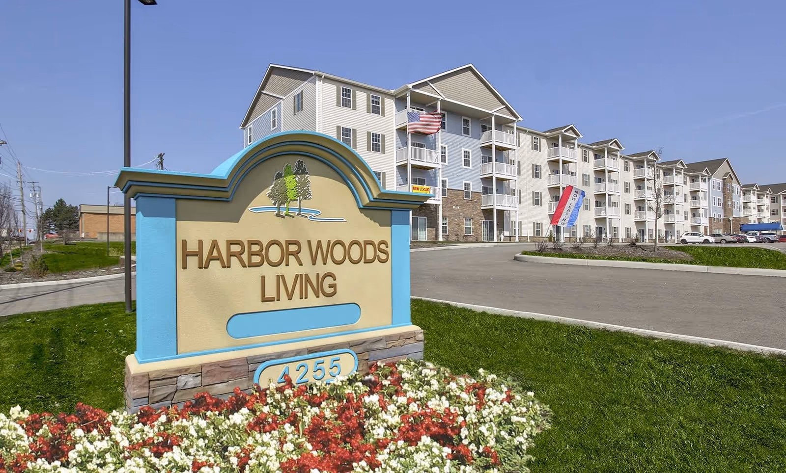 Entrance sign reading "HARBOR WOODS LIVING" in front of a multi-story senior living building, with flowers and a driveway.