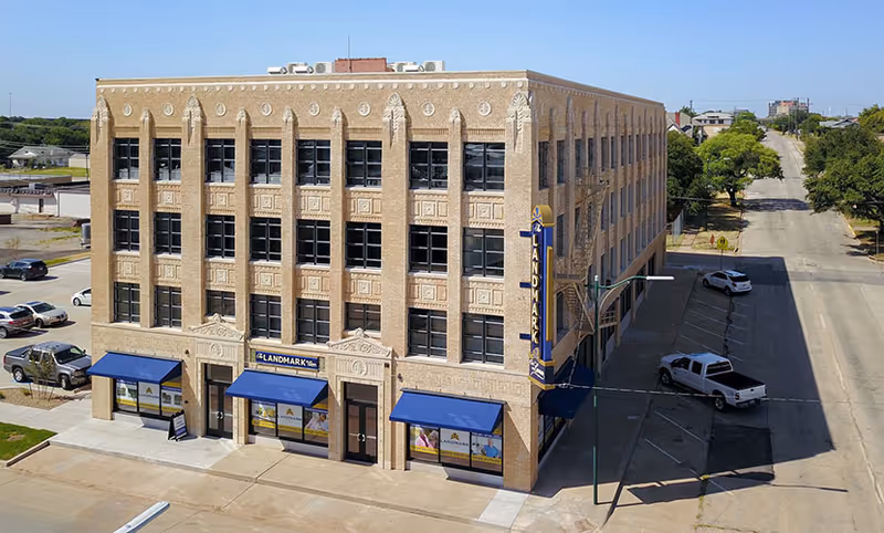 Front exterior of a four-story brick building labeled 'Landmark' with blue awnings and a vertical marquee sign on a corner lot.