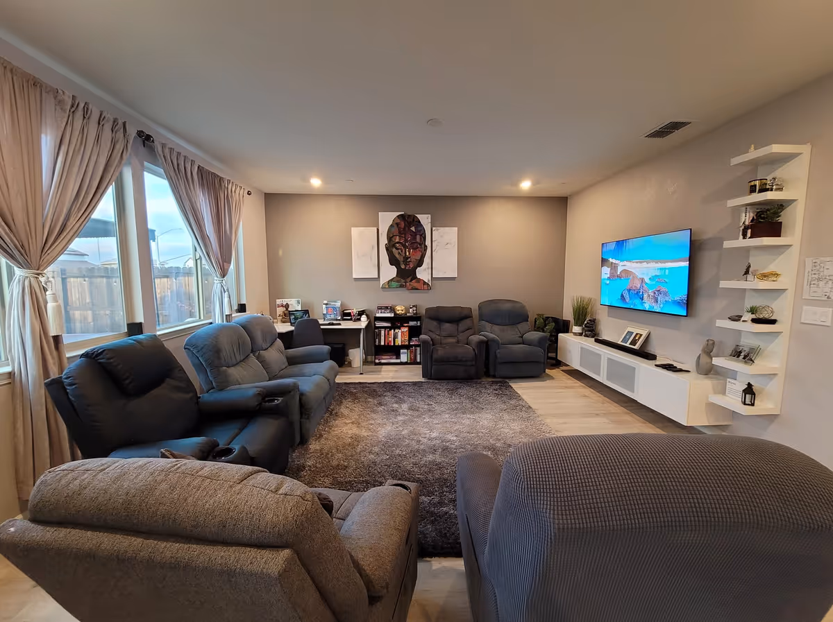 A cozy living room with multiple recliner chairs and sofas arranged around a large gray rug. The room has large windows with beige curtains on the left, a wall-mounted flat-screen TV on the right, and a small desk with a chair and bookshelf against the far wall. Decorative shelves with plants and ornaments are mounted on the right wall.