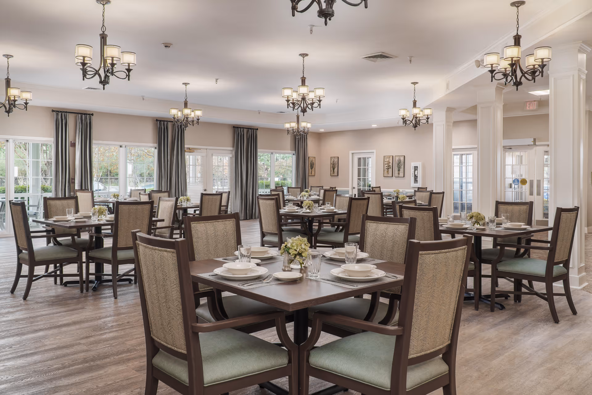 A spacious dining room with multiple square wooden tables, each set with white plates, bowls, glasses, and silverware. The chairs have wooden frames with green cushioned seats and patterned backs. The room has large windows with gray curtains, wooden flooring, and several elegant chandeliers hanging from the ceiling. There are decorative wall art pieces and a door leading outside.
