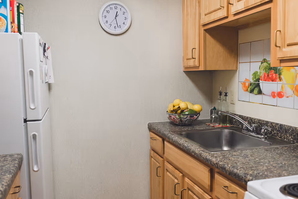 A small kitchen area with wooden cabinets, a granite countertop, a stainless steel sink, a white refrigerator, and a wall clock showing 12:05. There is a bowl of fruit on the counter and a decorative tile backsplash with images of vegetables.