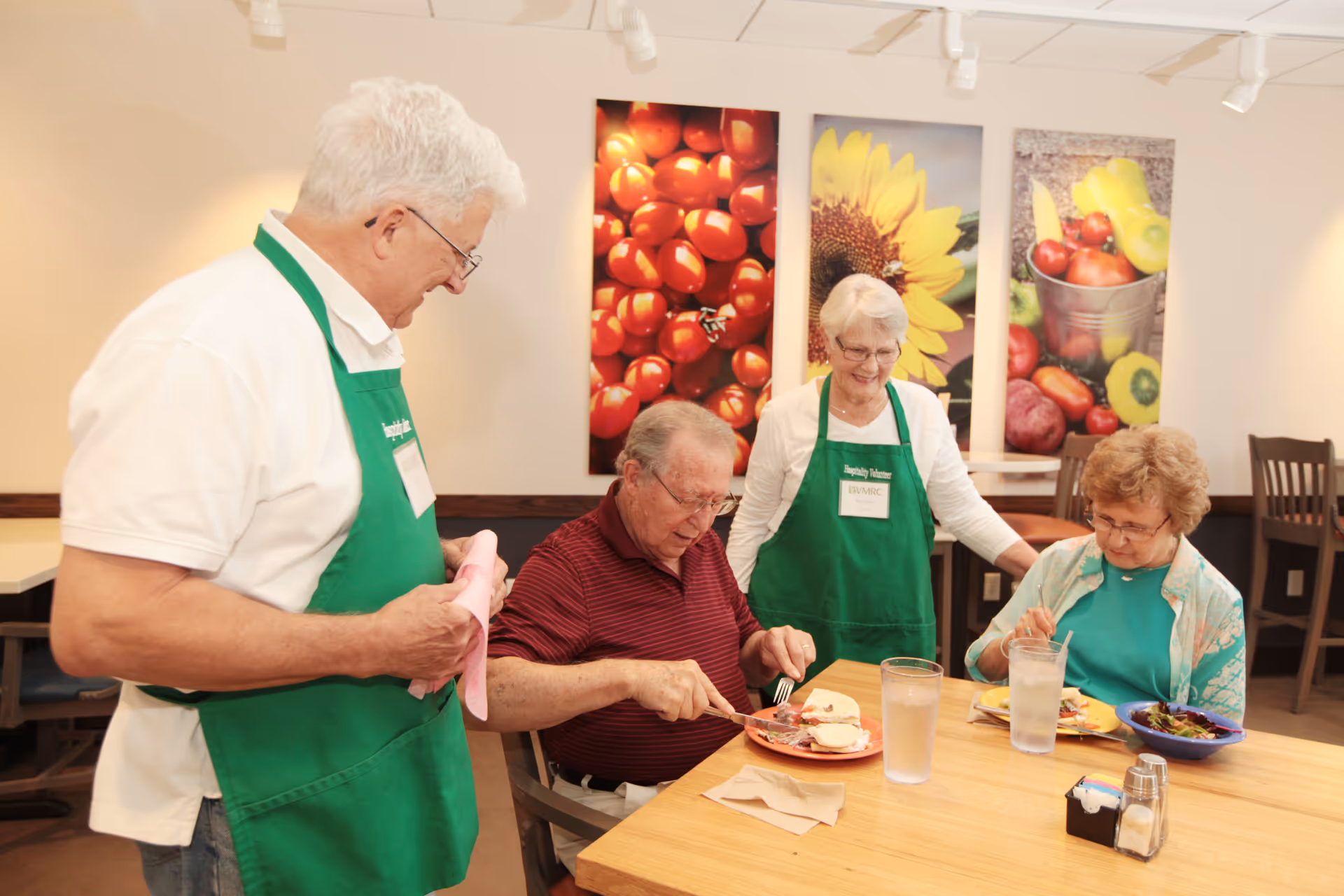 Three older adults and a volunteer in green aprons at a communal dining table with plates of food and colorful fruit-and-flower wall art behind them.