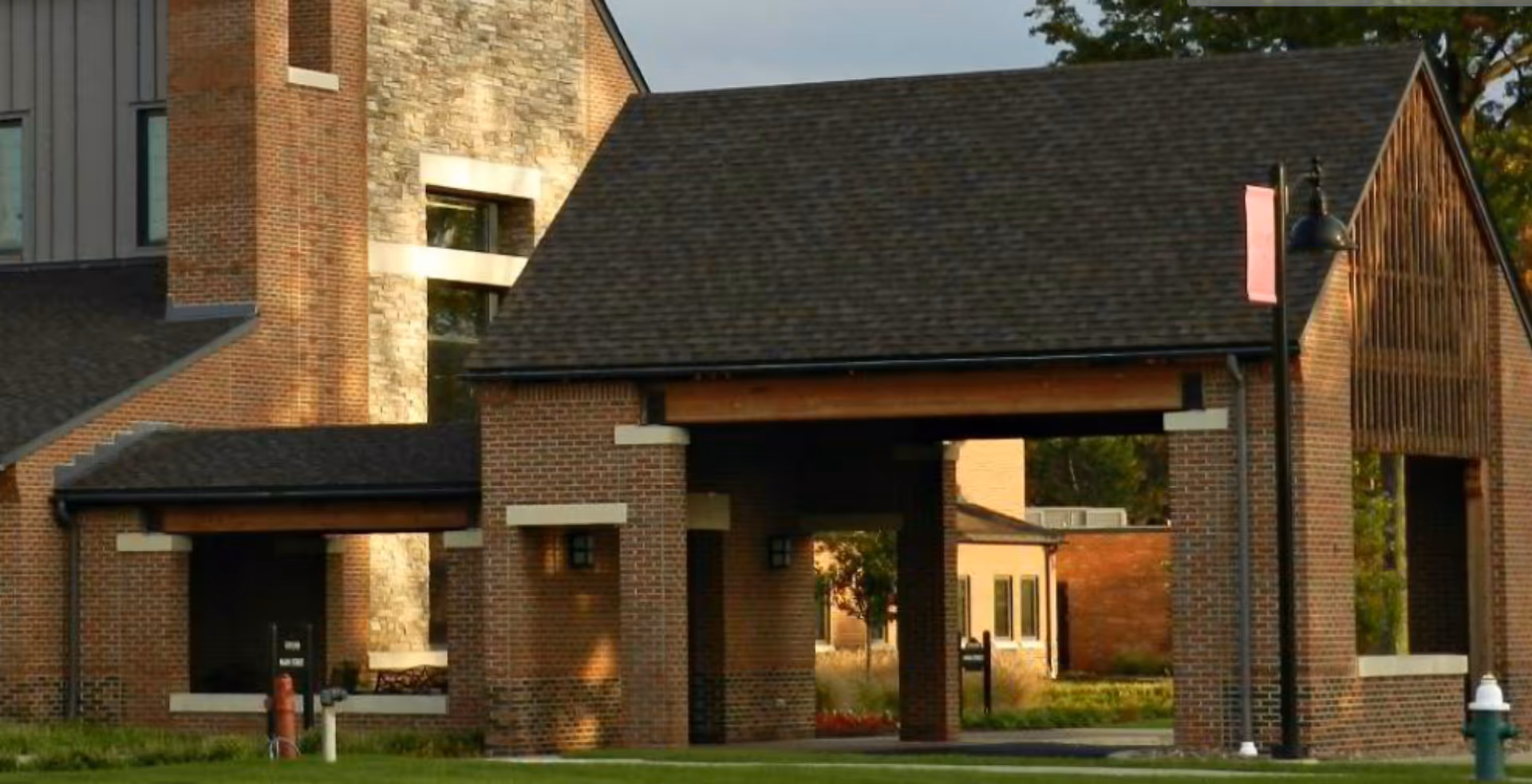 Exterior view of a brick building with a covered entrance supported by brick columns, featuring a shingled roof and a lamppost with a red banner nearby. The building has stone and brick walls and is surrounded by grass and trees.