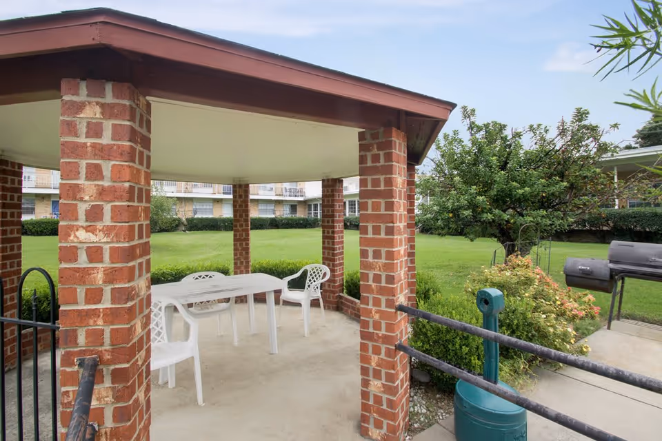 Covered brick gazebo with white plastic chairs and a table in a grassy courtyard of an apartment complex.