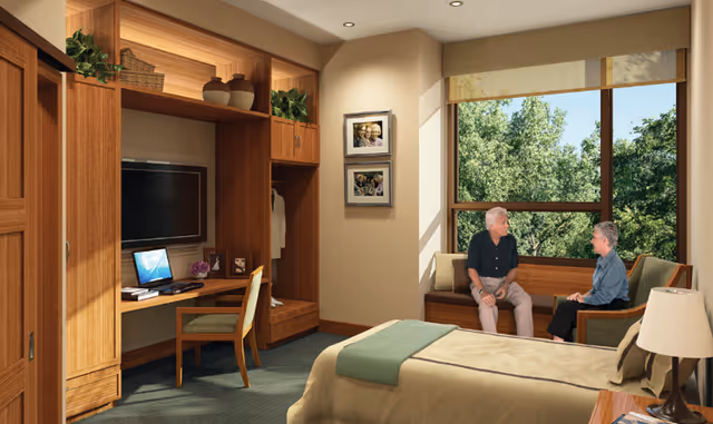 Sunlit bedroom with wooden built-in desk and cabinetry, bed in the foreground, and two people seated by a large window overlooking trees.