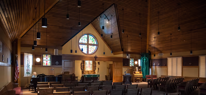Interior chapel with rows of chairs facing an altar, stained-glass windows, a piano, and a wood-paneled vaulted ceiling.