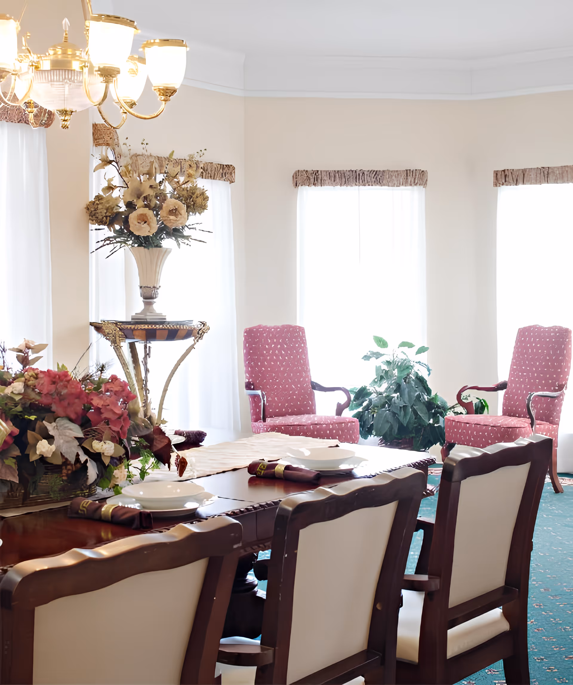 A bright dining room with a wooden dining table set with white plates and napkins. The room features upholstered chairs with wooden frames, two pink armchairs near large windows with sheer white curtains, and decorative floral arrangements on the table and a side table. A chandelier hangs above the dining table.