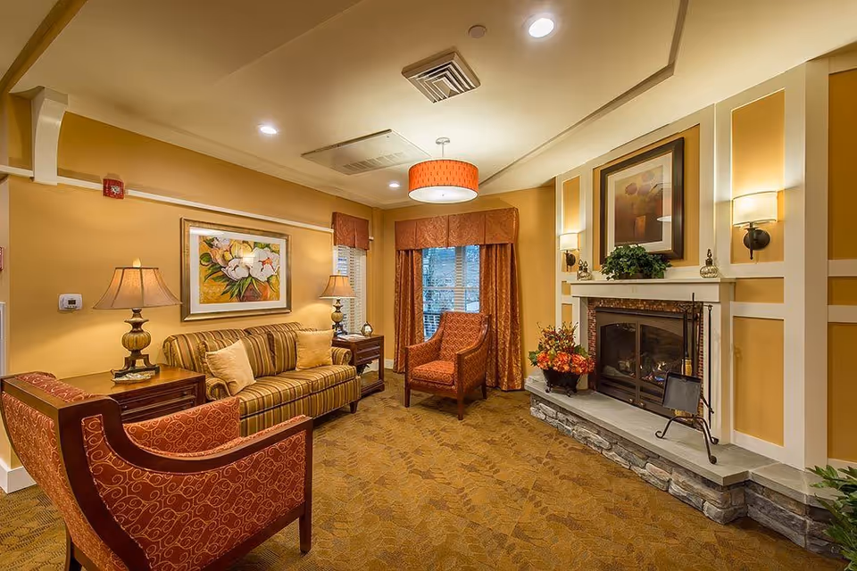 A cozy living room with warm yellow walls, a striped sofa with beige cushions, two patterned armchairs, a stone fireplace with a fire burning, framed artwork above the fireplace and sofa, two table lamps on side tables, and a window with orange curtains.