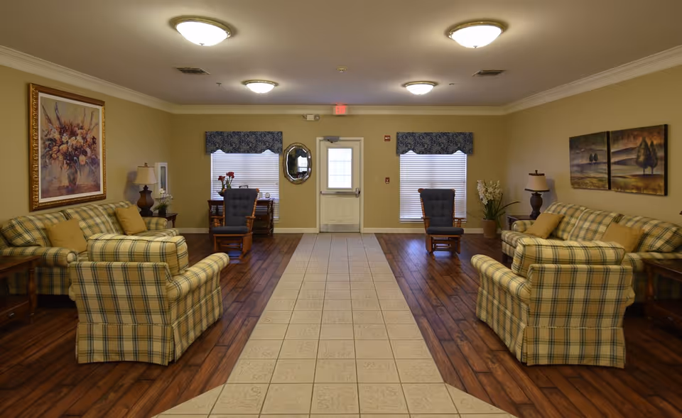 A spacious living room in Seguin Assisted Living featuring two plaid sofas and two matching armchairs arranged around a tiled walkway. The room has wooden flooring on the sides, beige walls adorned with framed paintings, two windows with blue valances, and ceiling lights. There are also two wooden rocking chairs near the windows and decorative plants and lamps on side tables.