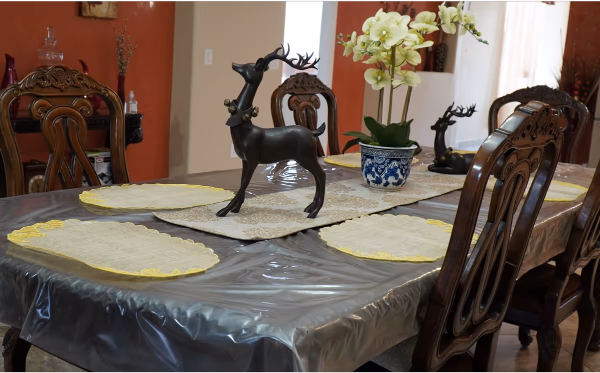 A dining table covered with a clear plastic tablecloth, set with four yellow-edged placemats and a decorative table runner. On the table are two black deer figurines and a blue and white ceramic pot holding white orchids. The table is surrounded by ornate wooden chairs, and the background shows a warm-toned wall with decorative items.