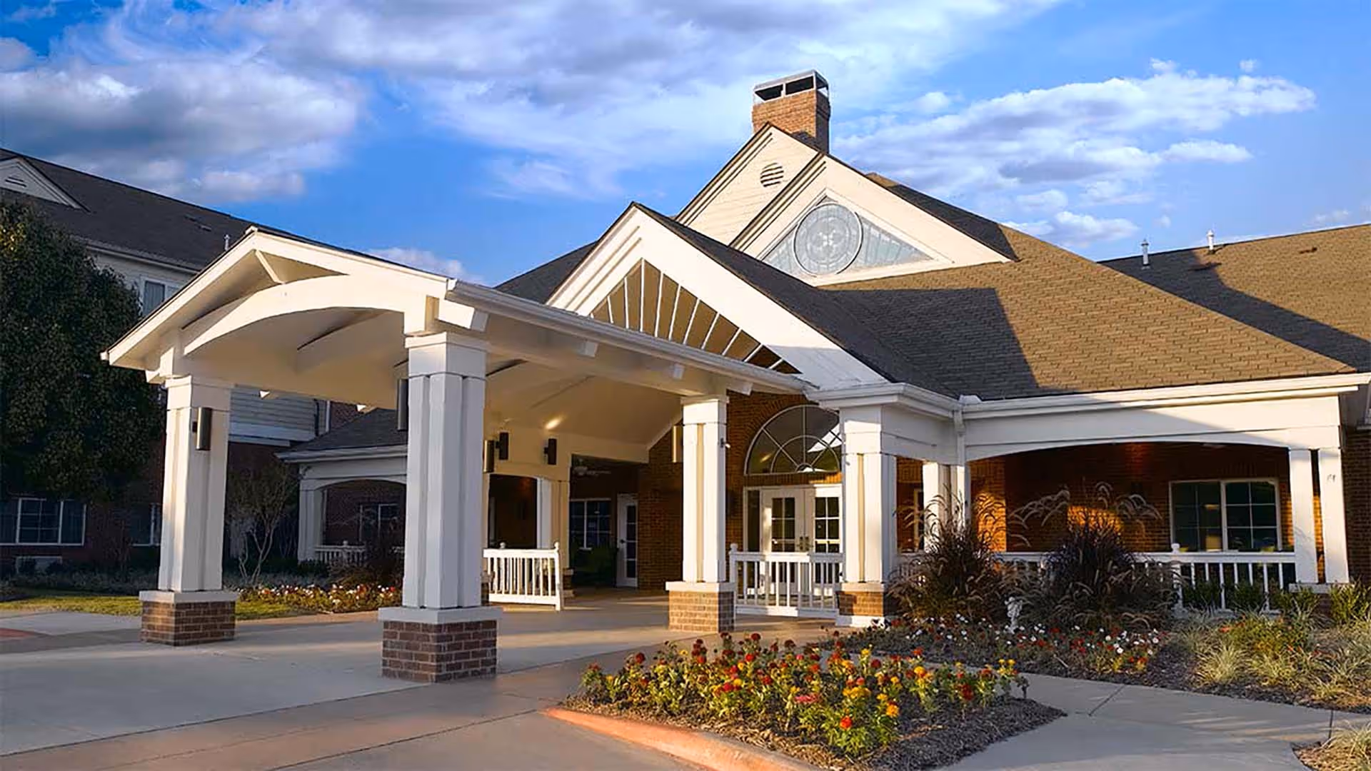 Exterior view of Atria Grapevine senior living facility showing the main entrance with a covered driveway, brick and white pillars, landscaped flower beds, and a partly cloudy sky.