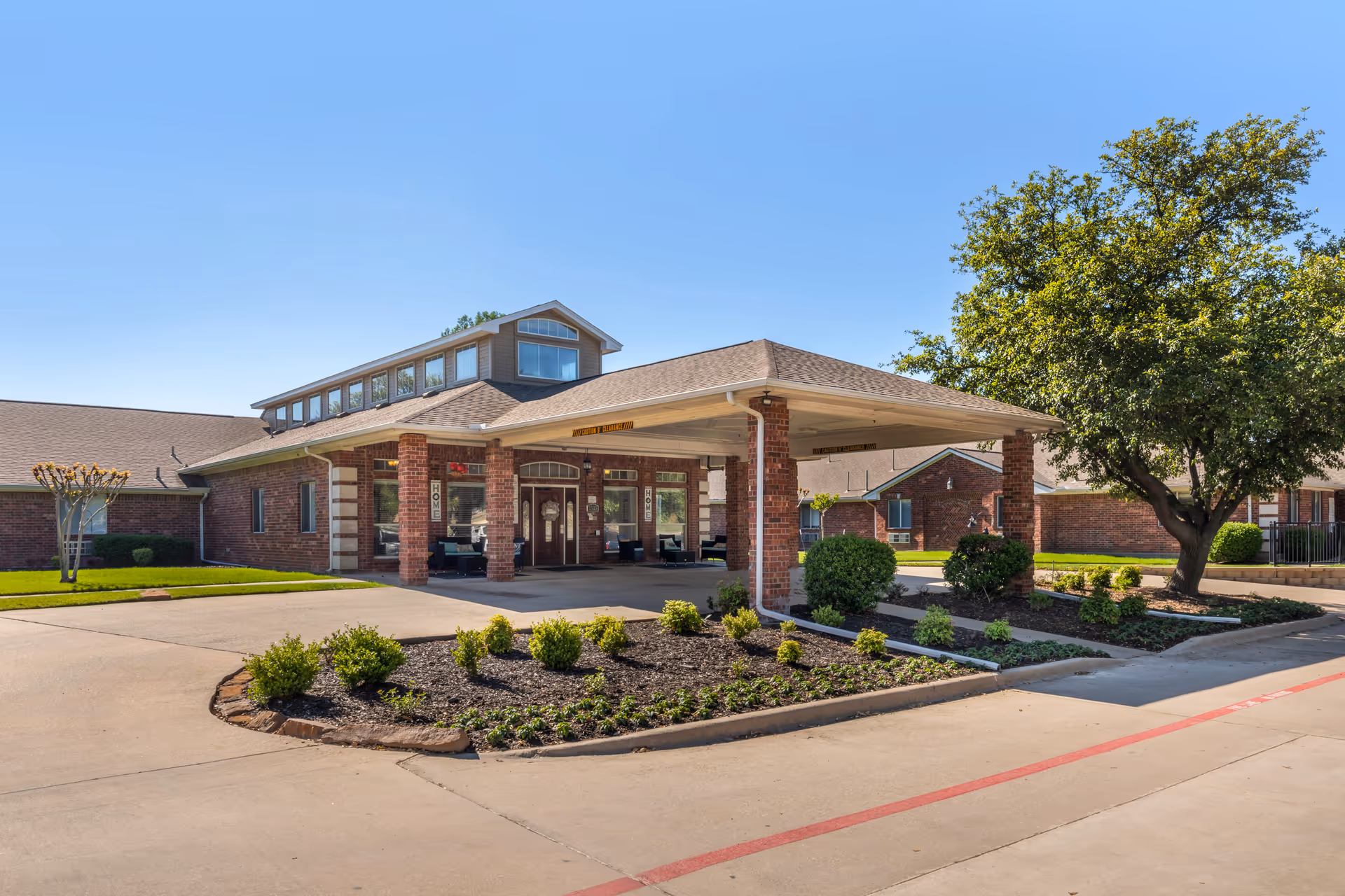 Exterior view of a senior living facility building with a covered entrance supported by brick pillars. There are landscaped bushes and a tree near the driveway, and the building has large windows and a sloped roof under a clear blue sky.