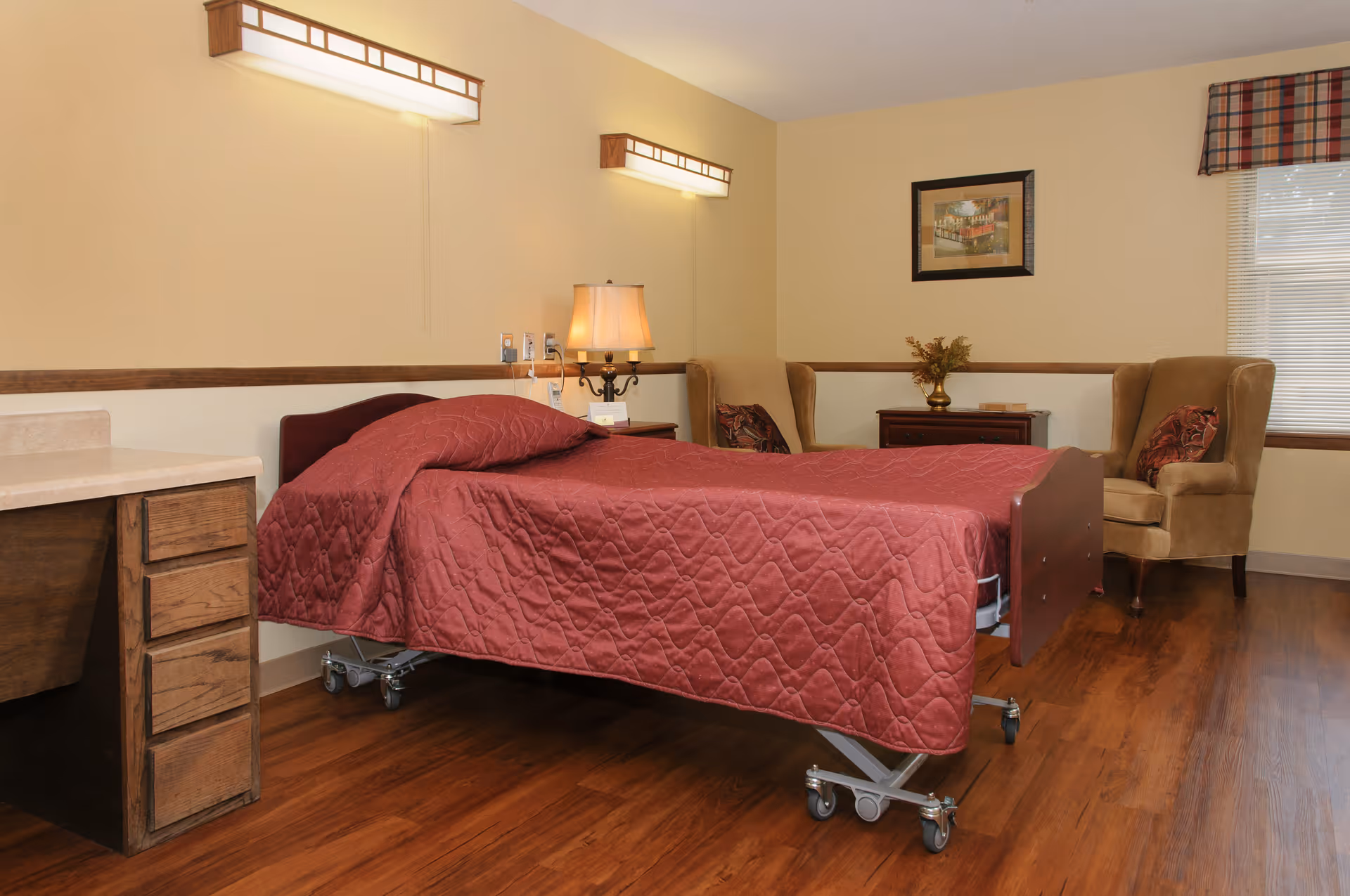 A senior living facility bedroom with a hospital-style bed covered in a red quilted blanket, a wooden desk with drawers, a bedside table with a lamp, two beige armchairs with decorative pillows, a framed picture on the wall, and a window with blinds and a plaid valance.
