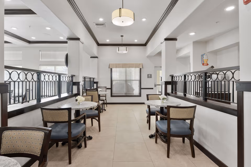 A bright and clean dining area with several round tables and cushioned chairs. The space features white walls with dark wood trim and railings, large windows with blinds, and modern ceiling lights. Small flower arrangements are placed on the tables.
