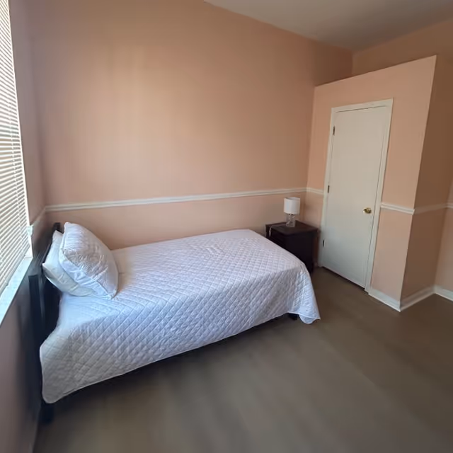 A small bedroom with peach-colored walls and a white door. There is a single bed with a white quilted bedspread and a pillow, positioned against the left wall near a window with blinds. Next to the bed is a dark wooden nightstand with a small white lamp on it. The floor is a light wood or laminate.
