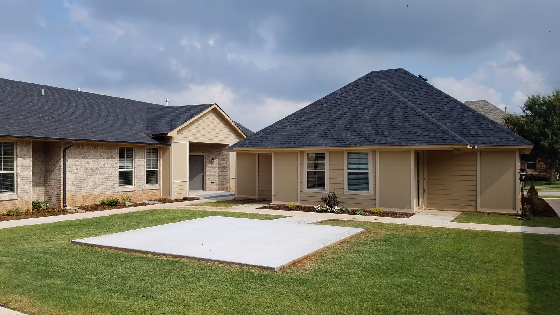 Exterior view of single-story residential buildings with beige siding and brick walls, dark shingled roofs, and several windows. The buildings are surrounded by green grass and concrete walkways under a cloudy sky.