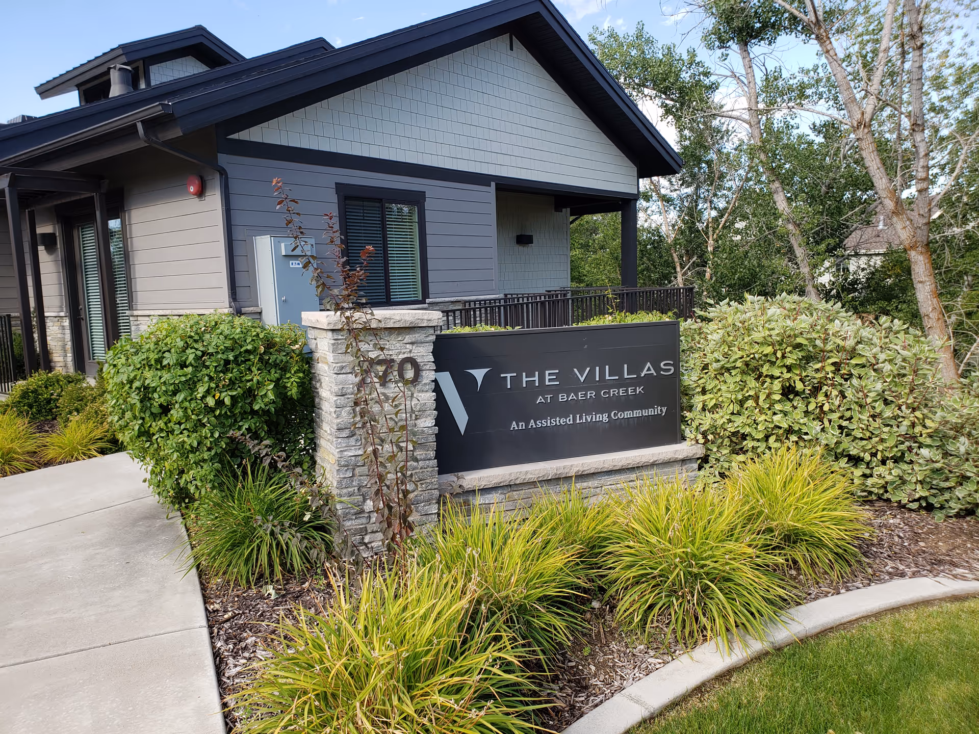 Exterior view of The Villas at Baer Creek assisted living community sign surrounded by green shrubs and plants with part of the building visible in the background under a partly cloudy sky.