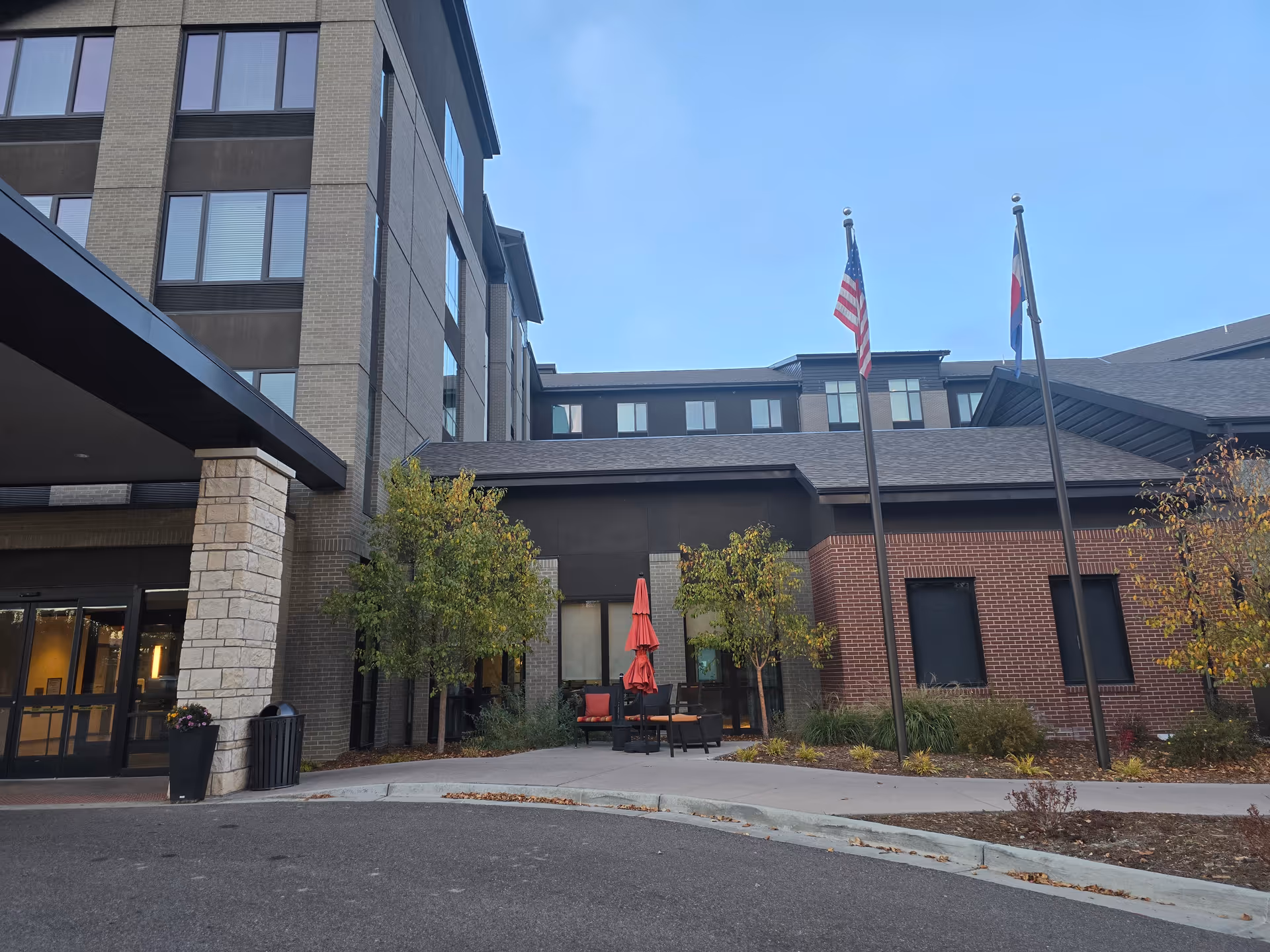 Exterior view of a senior living facility building with multiple floors, large windows, and a covered entrance. There are two flagpoles with the American flag and another flag, along with some outdoor seating with red umbrellas and trees near the entrance.