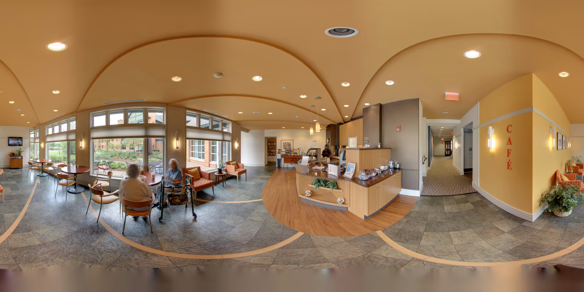 A bright and spacious café area inside The Summit at Brighton Independent Living facility, featuring large windows with natural light, several tables and chairs where two elderly individuals are seated and conversing, a reception counter with various items on display, and a hallway leading further into the building. The walls are painted in warm tones with the word 'CAFÉ' displayed prominently.