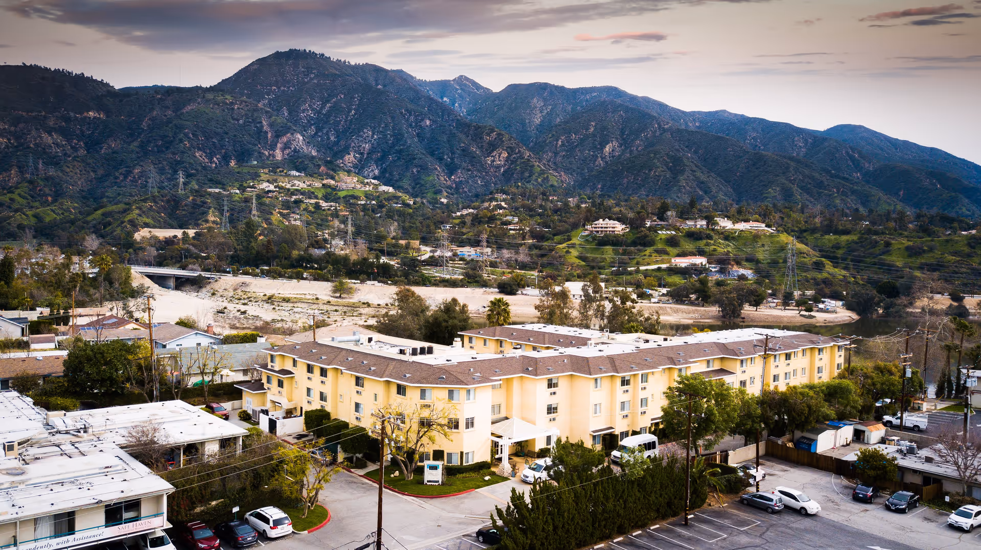 Aerial view of a large, multi-story yellow residential building surrounded by trees and parked cars, set against a backdrop of green hills and mountains under a cloudy sky.