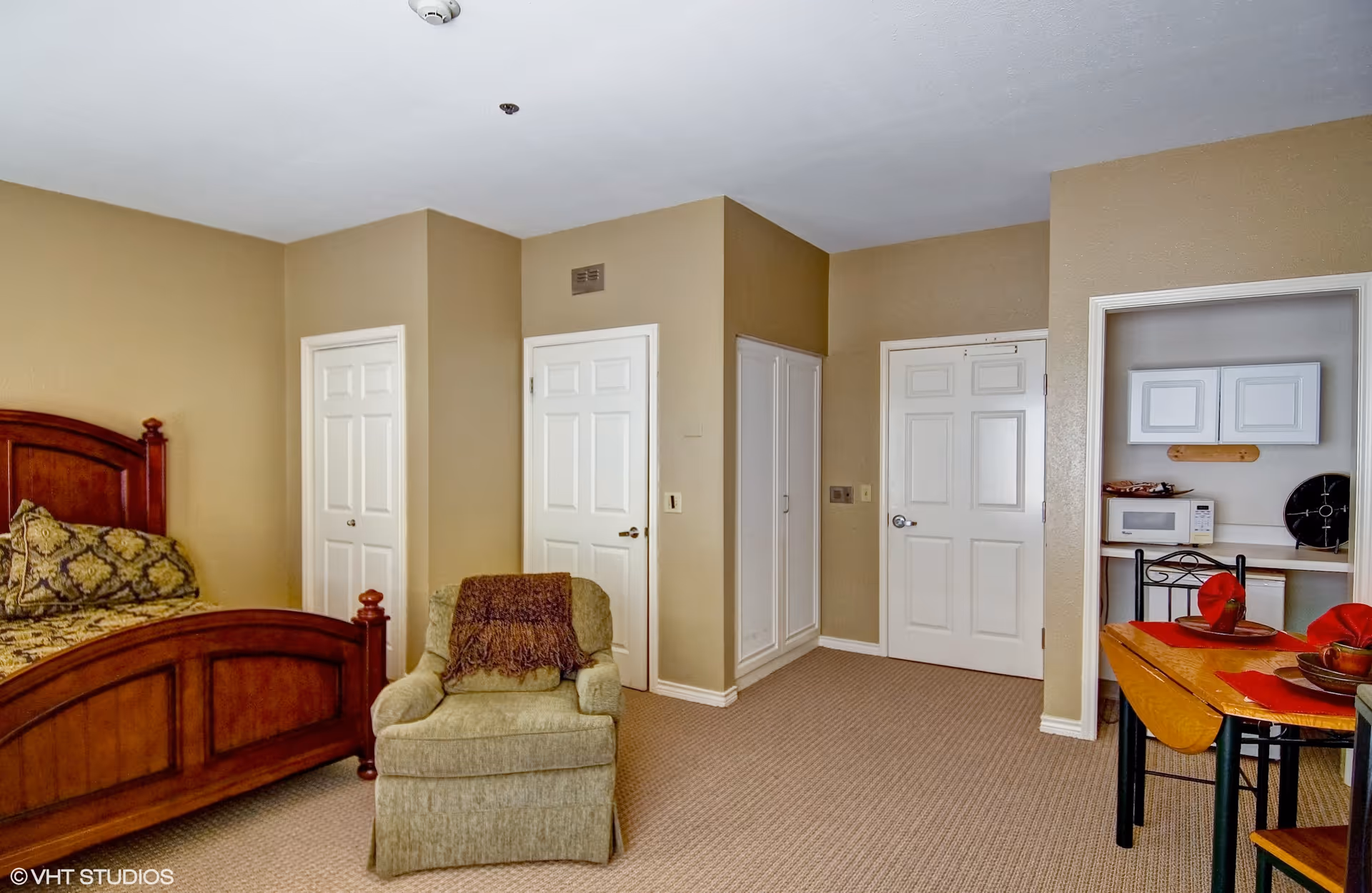 Interior view of a senior living facility room featuring a wooden bed with patterned bedding, a green upholstered armchair with a brown throw, beige walls, multiple white doors, and a small dining area with a table set with red napkins and a microwave on a counter in a small alcove.
