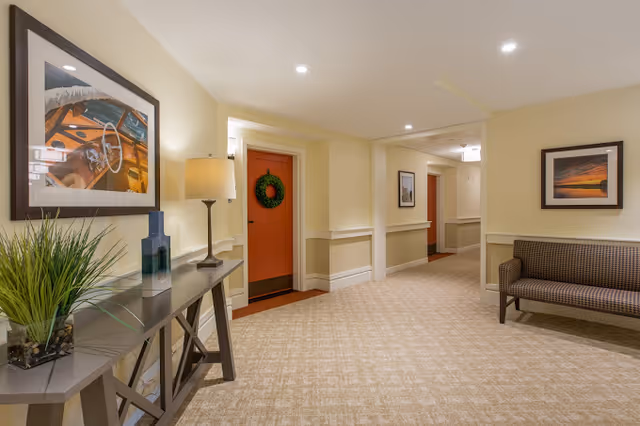 A well-lit interior hallway with a console table holding a lamp and plant, framed artwork, a seating bench, and doors (one decorated with a wreath).