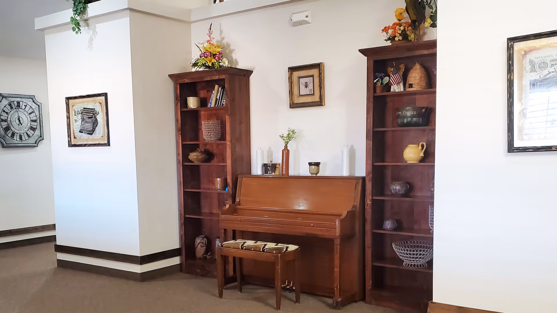 Upright wooden piano flanked by two tall wooden bookshelves and decorative wall art in a common room.