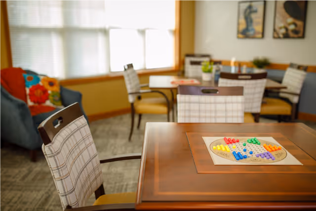 A cozy common area with several tables and cushioned chairs arranged around them. One table in the foreground has a colorful Chinese checkers board set up. The room has large windows with blinds, a blue armchair with colorful pillows, and framed artwork on the walls.