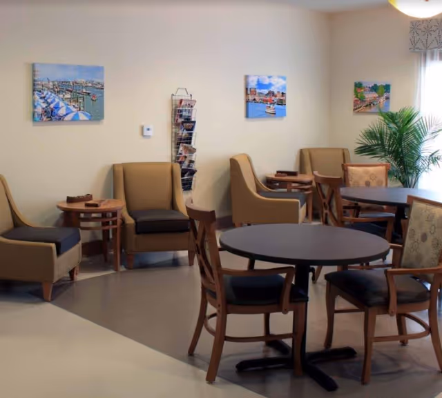 Communal dining and seating area with round tables, wooden chairs, upholstered armchairs, wall art, and a potted plant.