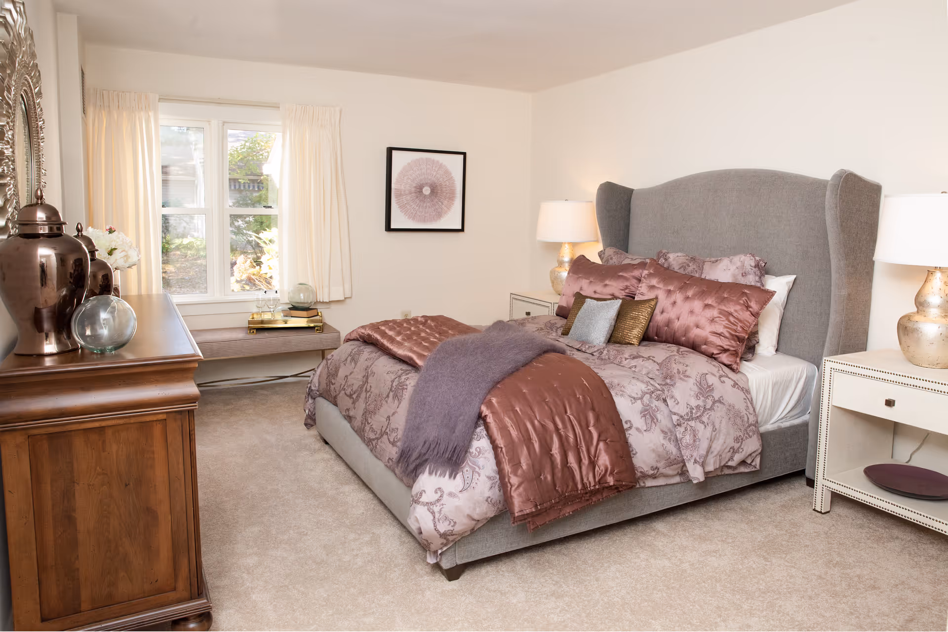 A nicely furnished bedroom with a grey upholstered bed, decorative pillows and throws, bedside tables with lamps, a dresser, and a window bench.
