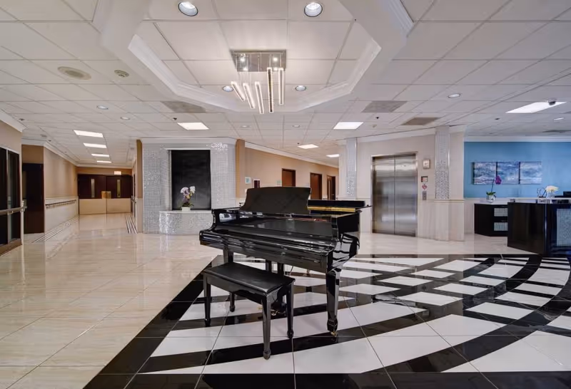 Spacious and well-lit lobby area with a black grand piano and matching bench placed on a stylish black and white tiled floor. The ceiling features recessed lighting and a modern chandelier. To the right, there is a reception desk with decorative artwork on the blue wall behind it. An elevator with closed doors is visible near the reception area. The hallway extends to the left with handrails along the walls.