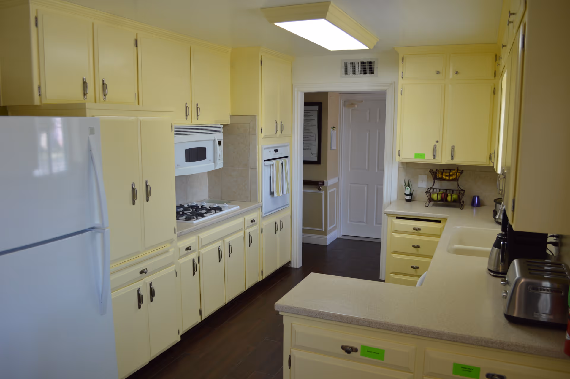 A bright kitchen with yellow cabinets and drawers, a white refrigerator, a built-in microwave above a gas stove, and a built-in oven. The kitchen has beige countertops with a toaster, a coffee maker, and a fruit basket on the counter. There is a doorway leading to another room with a white door and dark flooring.