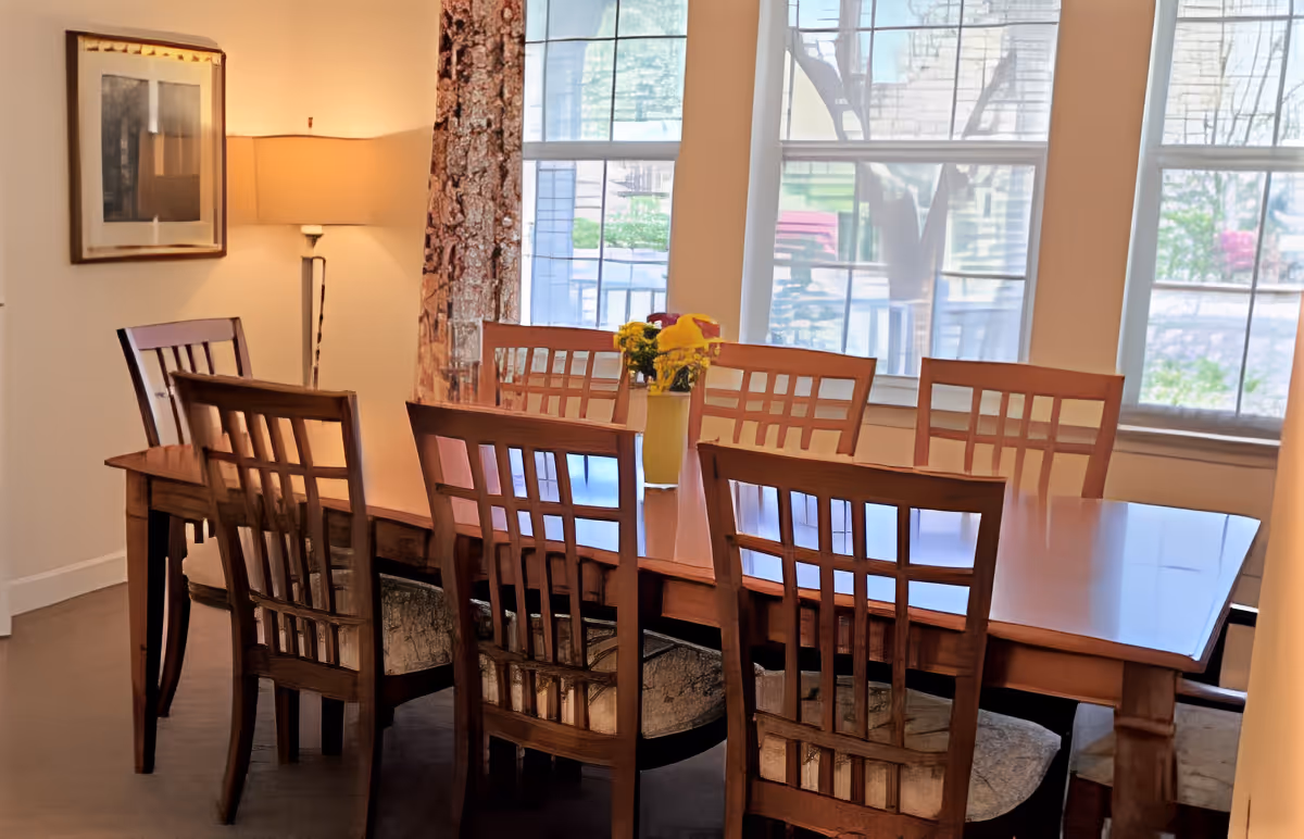 A dining room with a wooden table surrounded by eight wooden chairs with cushioned seats. A vase with yellow flowers is placed on the table. Large windows with white frames allow natural light to fill the room. A floor lamp and framed artwork are visible on the left wall.