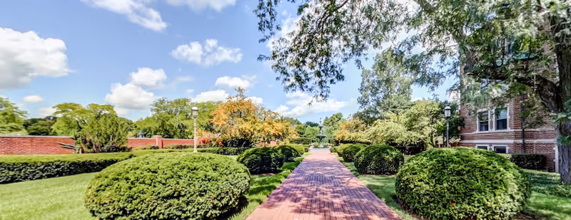 Brick walkway lined with trimmed round shrubs, trees, and lamp posts leading through landscaped grounds beside a brick building under a blue sky.
