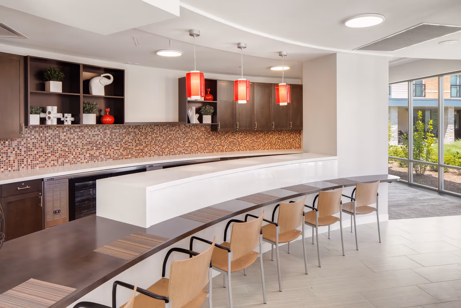 Modern kitchen area with a curved counter and six wooden chairs with metal frames. The backsplash features small mosaic tiles in shades of brown and red. Above the counter, there are three red pendant lights hanging from the ceiling. Dark wood cabinets and open shelves with decorative items are mounted on the wall. Large windows on the right side allow natural light to enter, showing some greenery outside.