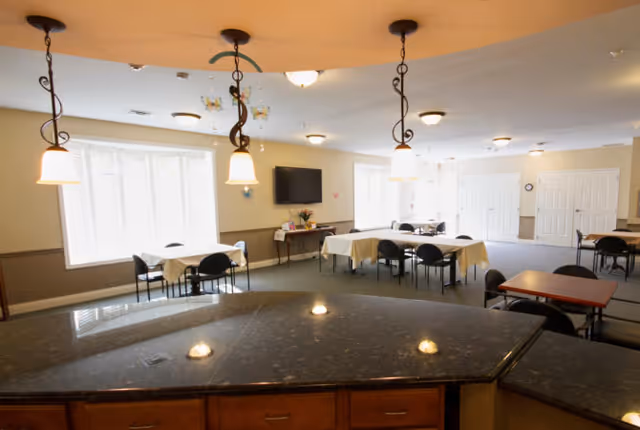 Interior view of a senior living facility common area with multiple tables covered with white tablecloths and black chairs around them. There are pendant lights hanging from the ceiling above a dark granite countertop in the foreground. The room has beige walls, large windows with blinds, a wall-mounted TV, and double doors in the background.