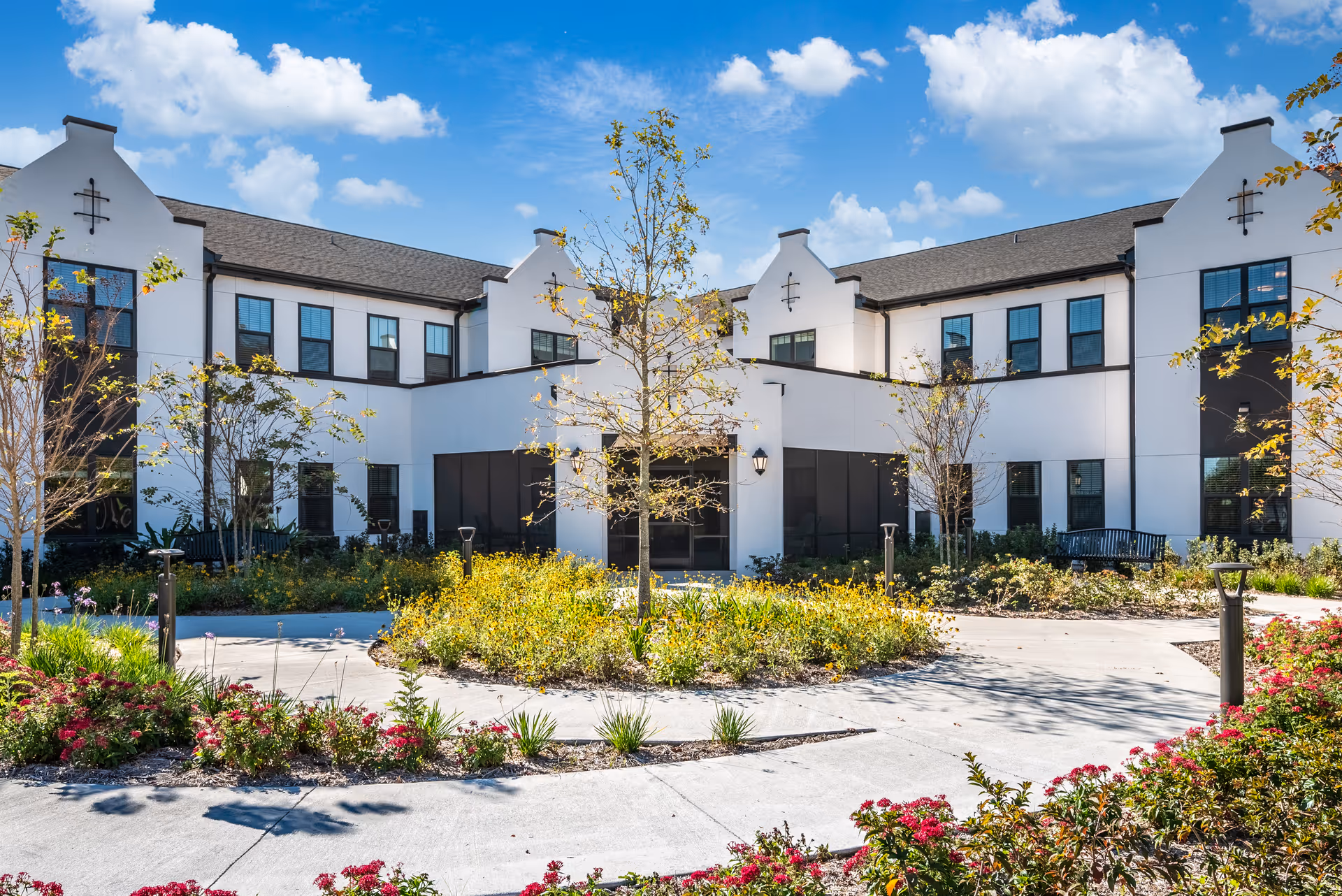 Exterior view of The Grove at Trelago senior living facility showing a two-story white building with black window frames, surrounded by landscaped gardens with trees, shrubs, and flowers under a blue sky with scattered clouds.