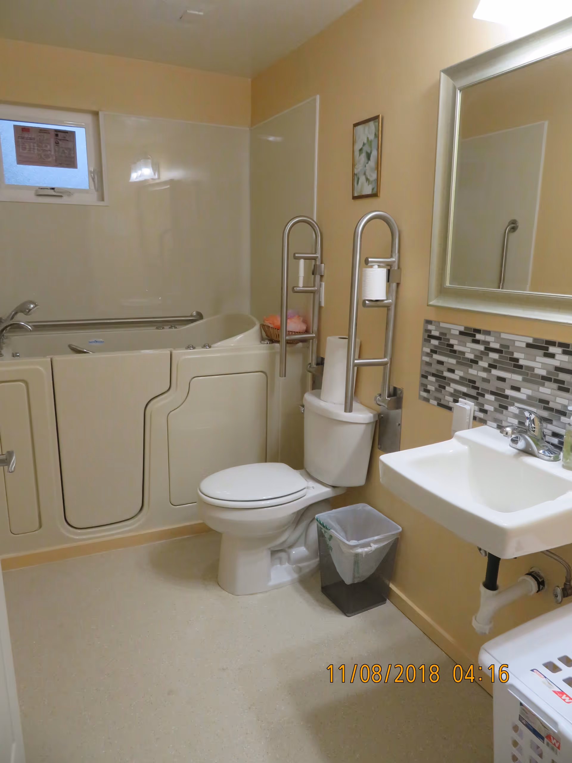 Handicap-accessible bathroom featuring a walk-in bathtub, toilet with grab bars, sink and mirror with a tiled backsplash.