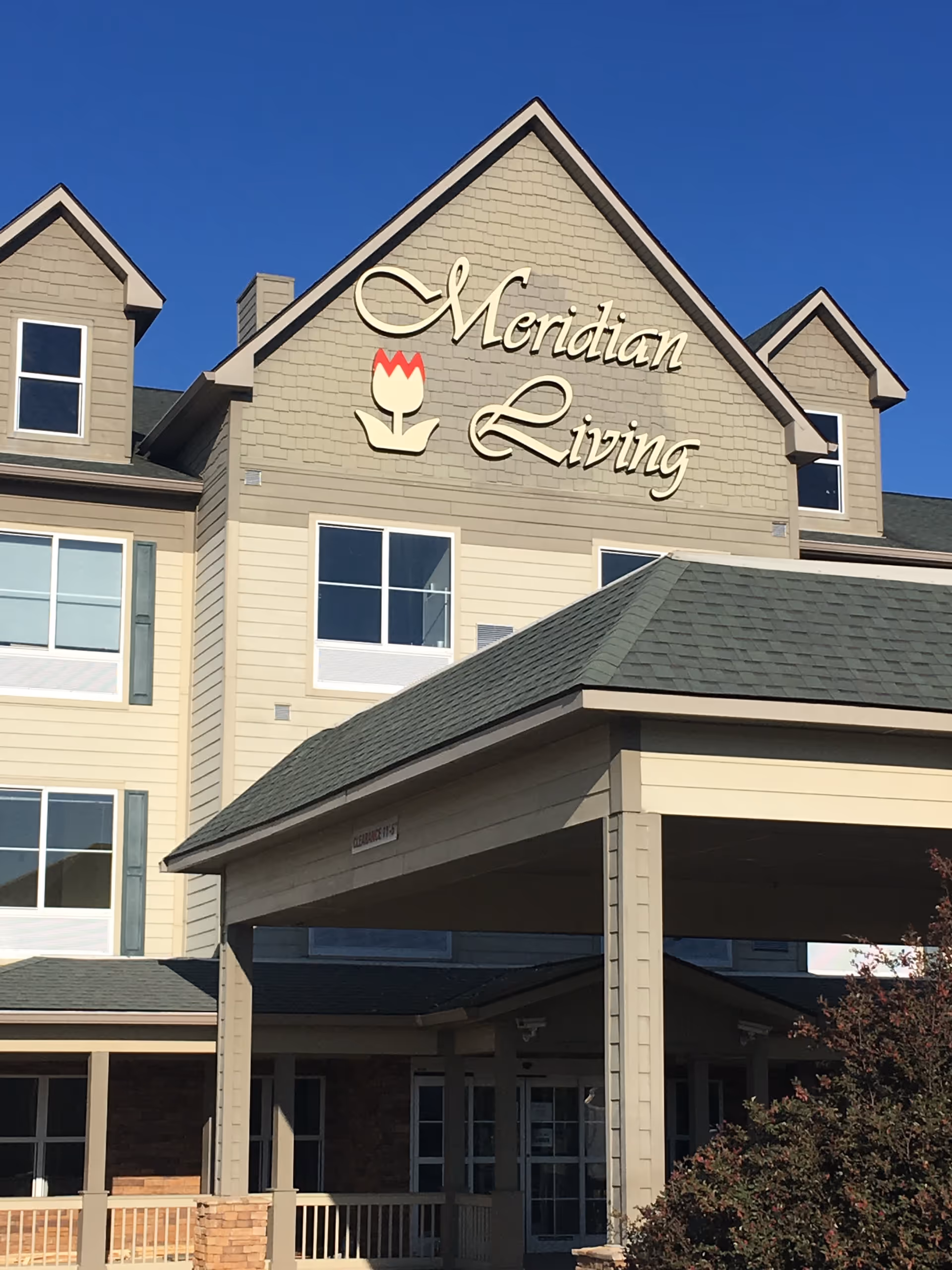 Exterior view of a multi-story building with beige siding and green roof shingles under a clear blue sky. The building has a covered entrance and a sign that reads 'Meridian Living' with a tulip logo above the entrance.