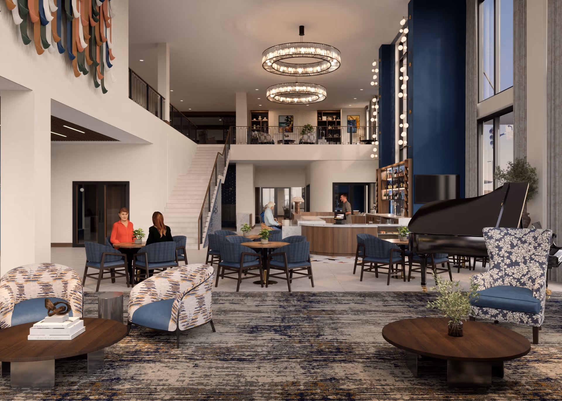 Spacious senior living facility common area with modern decor, featuring multiple seating arrangements including patterned armchairs and blue cushioned chairs around wooden tables. A grand piano is positioned near large windows with tall blue curtains. Two circular chandeliers hang from the ceiling. A staircase leads to an upper level with additional seating and bookshelves. Several people are seated and interacting in the space.