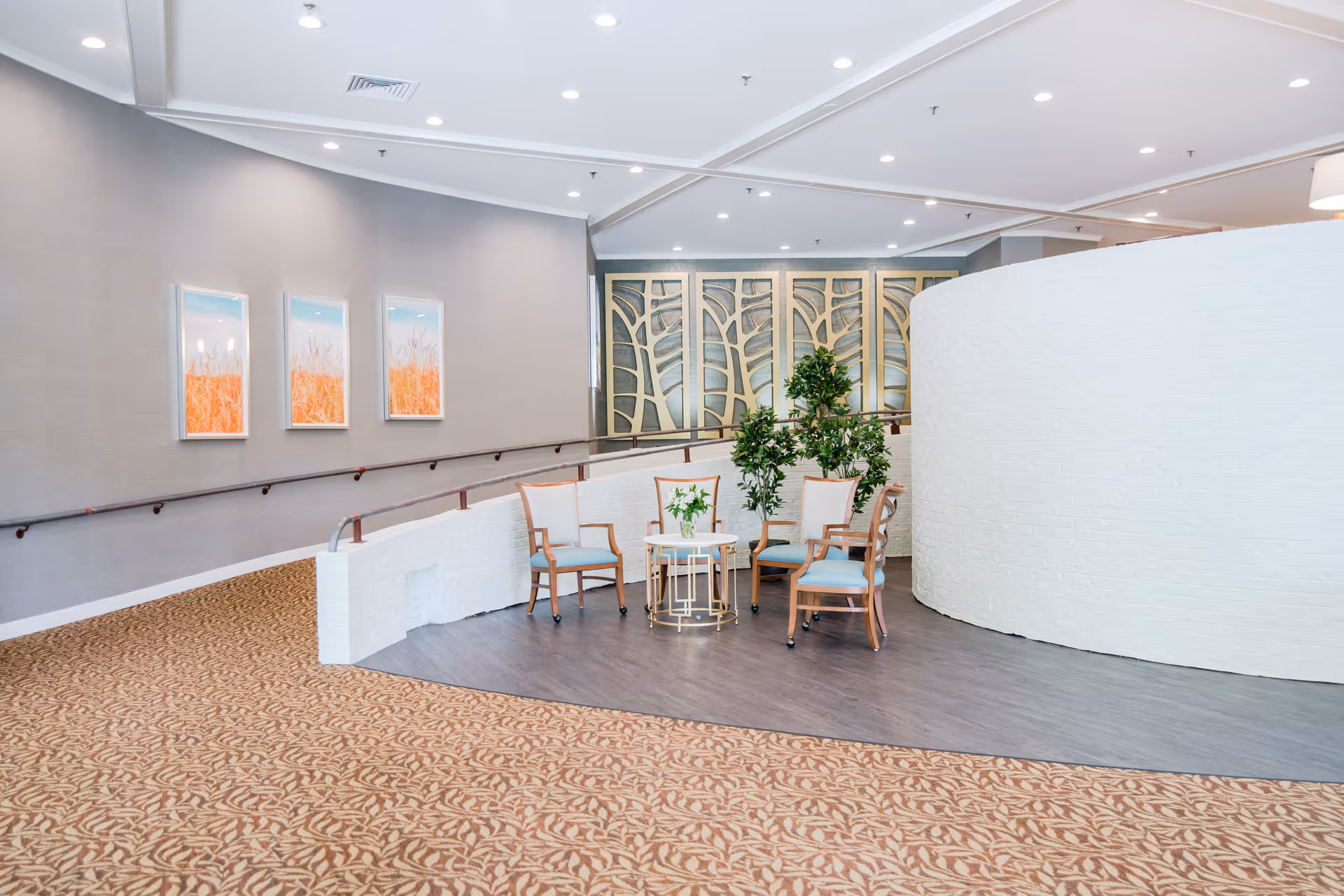 A bright, spacious interior seating area with four wooden chairs with light blue cushions arranged around a small round table with a flower vase. The area features a curved white brick wall, decorative wall panels with a tree branch design, and three framed pictures of orange wheat fields on a gray wall. The floor has a mix of patterned carpet and wood-like flooring, with ceiling lights illuminating the space.