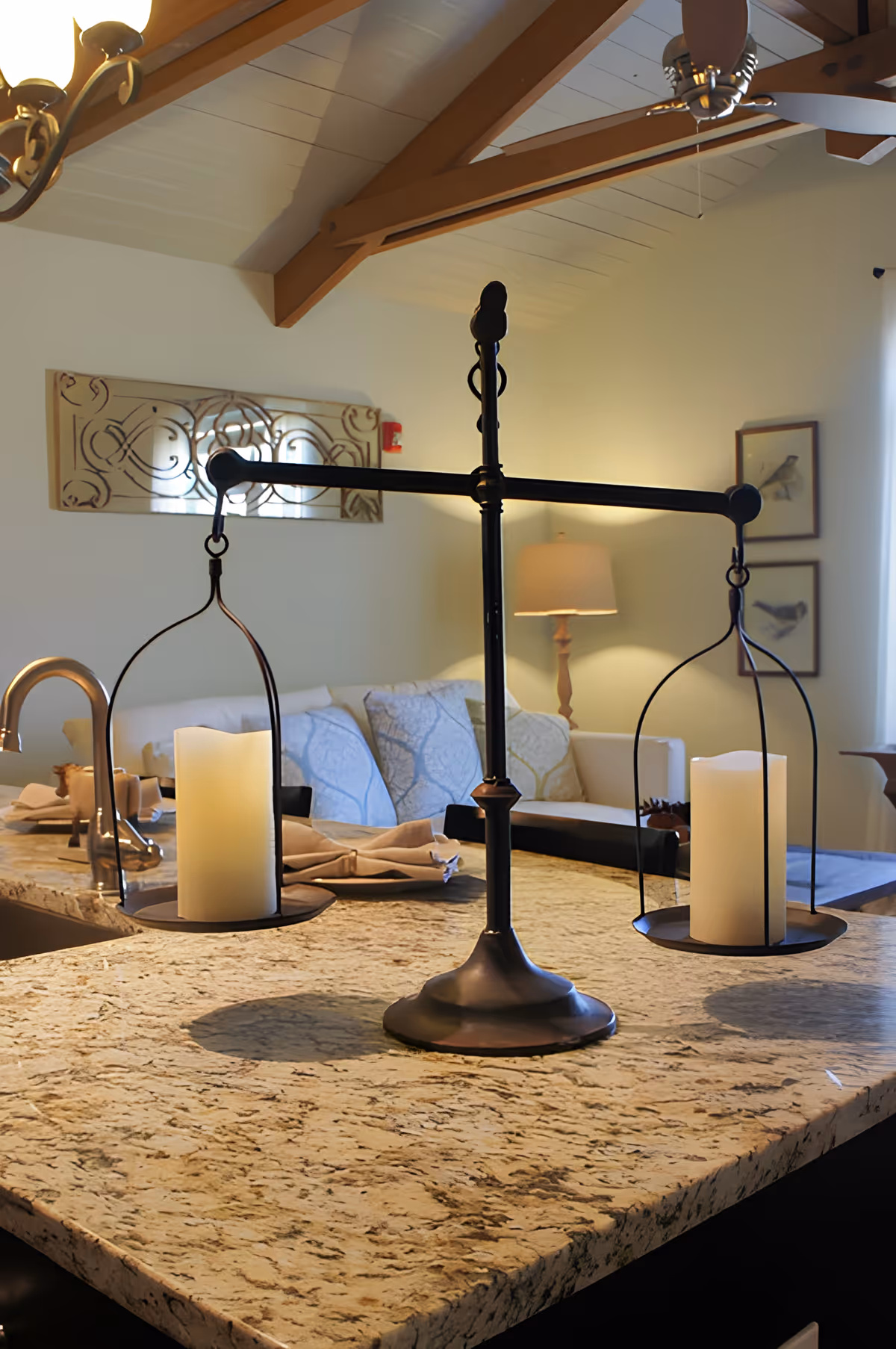 A cozy interior space featuring a granite countertop with a decorative balance scale holding two large candles. In the background, there is a white couch with patterned cushions, a floor lamp, framed bird artwork on the wall, and exposed wooden ceiling beams with a ceiling fan.