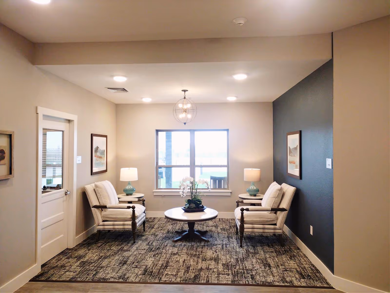 A cozy sitting area with two cushioned armchairs facing each other, each accompanied by a small round side table with a lamp. A round coffee table with a flower arrangement is centered between the chairs. The room has a window letting in natural light, neutral-colored walls with one dark accent wall, and framed artwork on the walls.